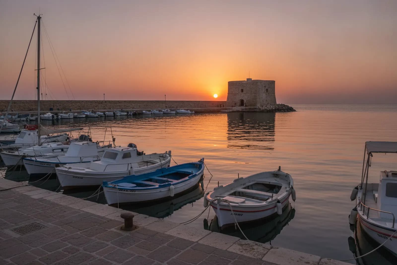 Piccolo forte di calcare in fondo al molo di un porto al tramonto, con le barche ormeggiate riflesse nell’acqua ferma