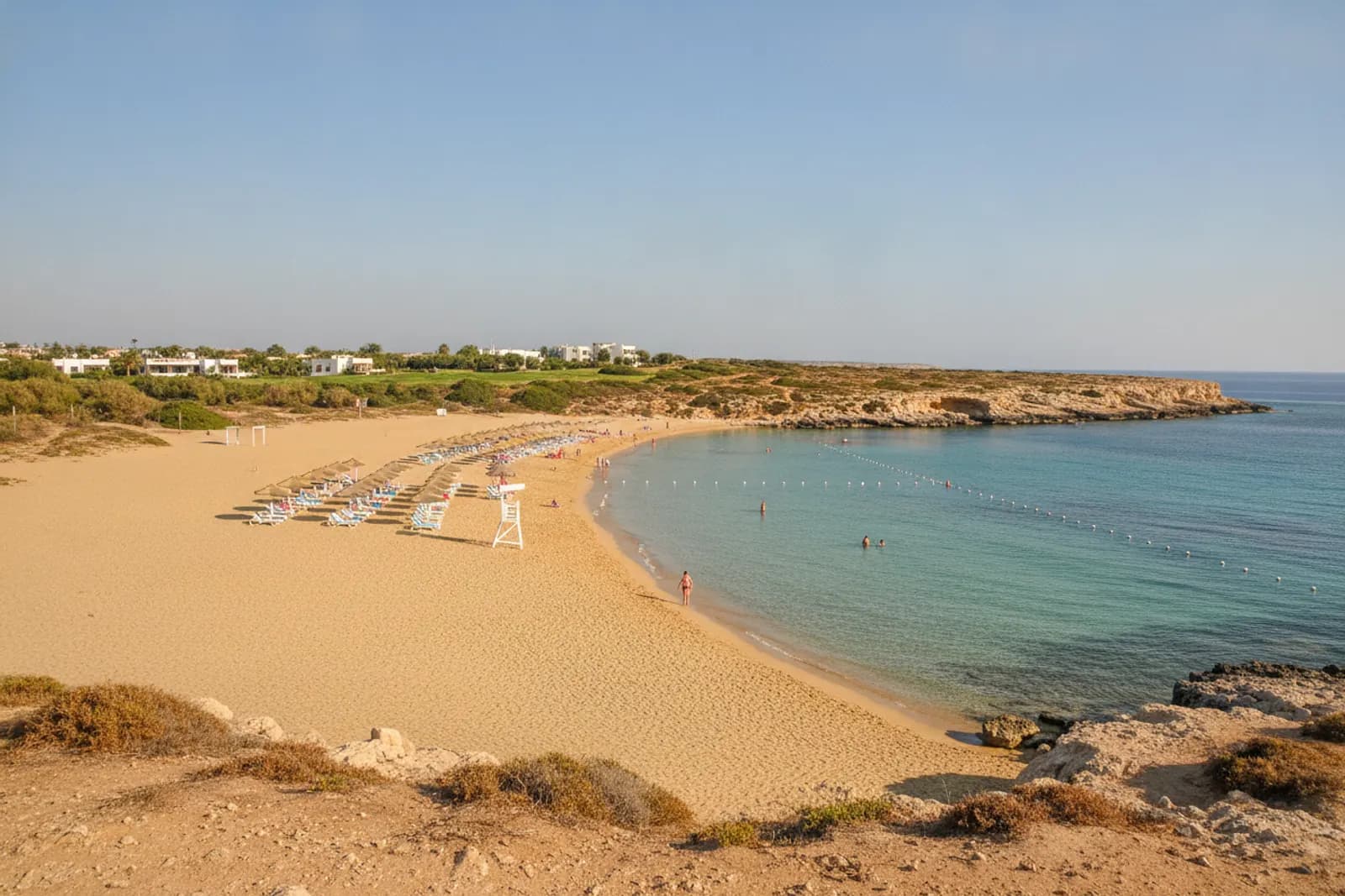 Baia ad arco di sabbia dorata e acqua turchese bassa sotto un basso promontorio calcareo, con lettini e ombrelloni